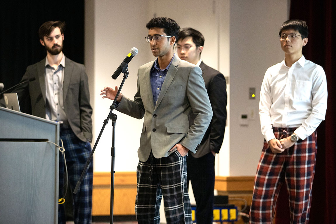 Four college students wearing blazers and plaid pajama pants on stage; one speaks into a microphone